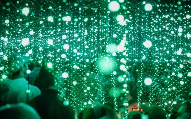 An abstract mesh of photographs overlayed on top of each other. Hundreds of LED strands take up the frame, some in focus some not. Audience members are blurred and illuminated light green.