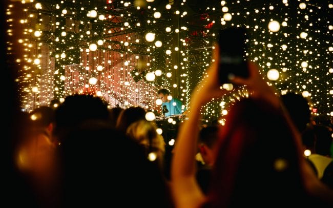 Four Tet is partially visible through hanging strands of LED lights. Audience members stand close together in front of him, illuminated in pink and yellow. A person in the foreground holds their phone above their head to take a photograph.